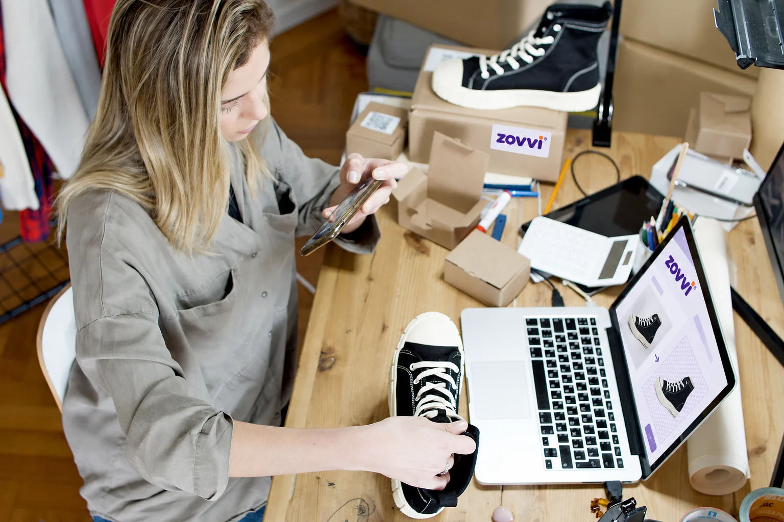 Comerciante fotografiando una zapatilla con el móvil junto al portátil y los paquetes de pedidos